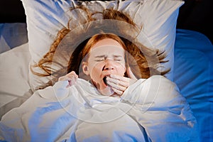 View top. a woman with red hair lies in bed on white bed linen and yawns.