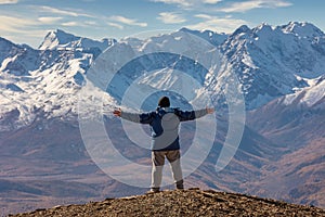 View of a tourist spreading his arms and facing the mountains. White snowy mountain ridge and beautiful blue cloudy sky as a