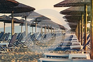 View of tropical beach with big straw umbrellas and sun loungers on the sunset sea background