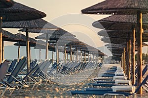 View of tropical beach with big straw umbrellas and sun loungers on the sunset sea background