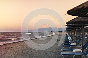 View of tropical beach with big straw umbrellas and sun loungers on the sunset sea background