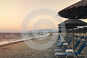 View of tropical beach with big straw umbrellas and sun loungers on the sunset sea background