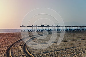 View of tropical beach with big straw umbrellas and sun loungers on the sunset sea background