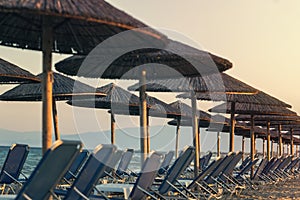 View of tropical beach with big straw umbrellas and sun loungers on the sunset sea background