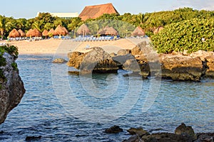 View of a tropical sandy beach with straw umbrellas, lounge green vegetation and coral reef rocks.