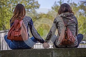 View of two girlfriends girls sitting on a wall and looking at the landscape hand in hand and lovingly