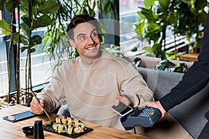 View of waitress holding payment terminal near cheerful man with smartphone