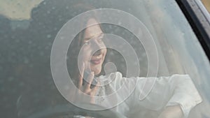 View through wet with rain windscreen of smiling girl in car talking on phone