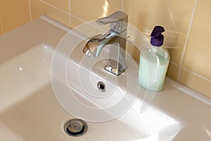 View of a white bathroom sink with a contemporary mixer tap, an unlabeled bottle of liquid soap. Concept of modern interior, water
