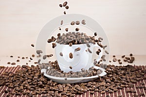 View of white cup standing on white plate with roasted coffee beans on tablemat.