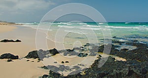 View of a white sand beach with black rocks. Blue transparent ocean waves splashing foam. Shoreline of paradise tourist