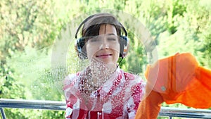 View through the window, smiling woman in headphones, in gloves, Cleaning Window by spraying Cleaning Products, using