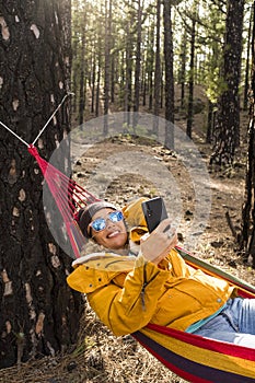 View of woman enjoying freedom and independence lay down on colorful hammock in the forest woods nature park and using modern