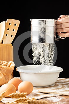 View of woman`s hand with flour falling from a sieve into a bowl, on a rustic table with cloth, eggs, ears of wheat and wooden sp