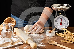 View of woman`s hand on pizza dough, falling flour, on wooden table with rustic kitchen objects, black background,