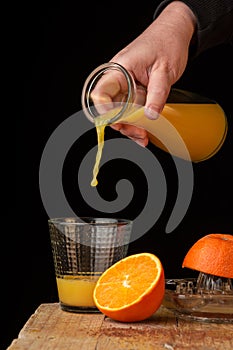 View of woman`s hand serving frozen orange juice with a bottle, over a glass, on wooden table with oranges, black background