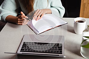 View of woman studying online with notepad and laptop in cafe with cup of coffee