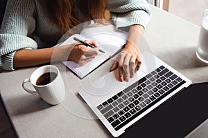 View of woman writing in notepad and using laptop in cafe with cup of coffee