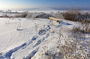 View of a wooden bench standing on a high bank of the river on a bright winter day. Rural winter landscape