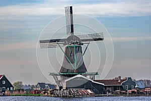 View of a working windmill in the middle of an old Zaans village on a river. background a striped blue and white sky.