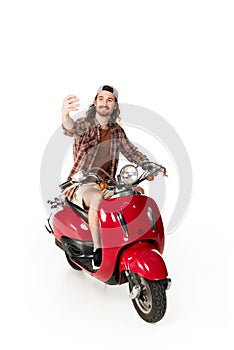 View of young man sitting on red scooter and taking selfie isolated on white