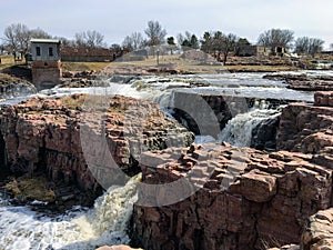 The Big Sioux River flows over rocks in Sioux Falls South Dakota with views of wildlife, ruins, park paths, train track bridge, tr