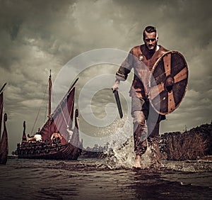 Viking warrior in the attack, running along the shore with Drakkar on the background.