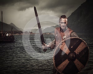 Viking warrior in the attack, standing along the shore with Drakkar and mountains on the background. photo