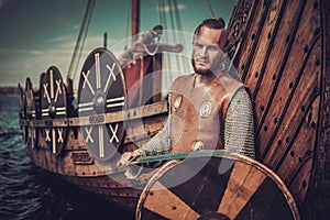 Viking warrior with sword and shield standing near Drakkar on the seashore.