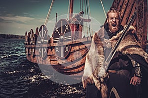 Viking warrior with sword and shield standing near Drakkar on the seashore.