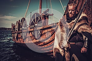 Viking warrior with sword and shield standing near Drakkar on the seashore.