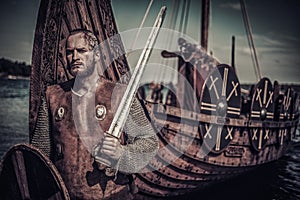 Viking warrior with sword and shield standing near Drakkar on seashore.