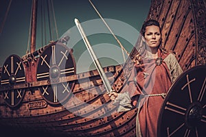Viking woman with sword and shield standing near Drakkar on the seashore.