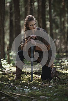 Viking woman with sword wearing traditional warrior clothes in a deep mysterious forest.