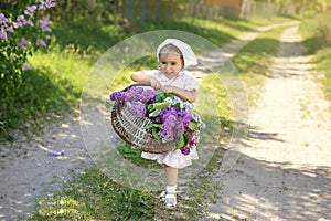 A village girl in a white dress and a kerchief carries a basket filled with spring blooming flowers and violet lilac along a count