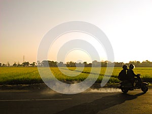 Village landscape captured during sunrise, motorbike rides crossing rural roads, sunlight peering through fog and trees on the gro