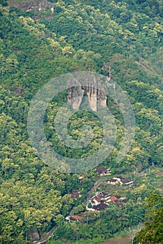 A village nestled at the base of a verdant hill, dotted with large rock formations that cling to the slope.