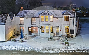 A village pub at night in the snow seen from above. The light are on and the glow looks warm and welcoming