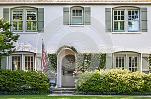 Vine covered entrance to white painted brick house with arched front door and wreath and arched windows with green shutters - land