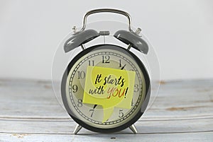 Vintage alarm clock with yellow speech bubble written with inspirational motivation quotes It Starts With You