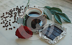 Vintage beautiful cup of hot coffee, bright red rose, green leaves, scattered coffee beans and dark chocolate bar on a wooden tabl