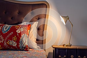 Vintage bed corner with a brown headboard, antique style pillows, and a bedside table with a lamp