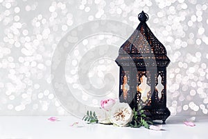 Vintage black Moroccan, Arabic lantern with glowing candle, green branches, rose flowers and petals on white table