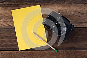 Vintage camera with empty yellow note and pencil on wooden background