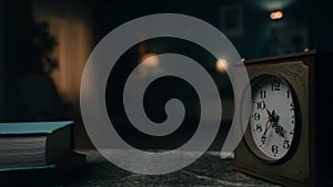 A vintage clock stands on a table against a blurred background of a room plunged into darkness close up. Time is running