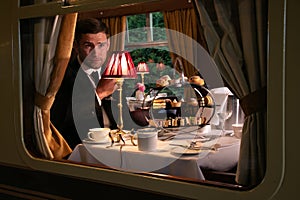 Vintage, handsome male in suit enjoying afternoon tea in train carriage with cakes, sandwiches and tea