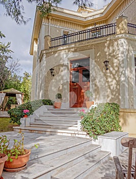 A vintage house entrance with marble stairs to the natural wooden double doors and potted plants through the garden.