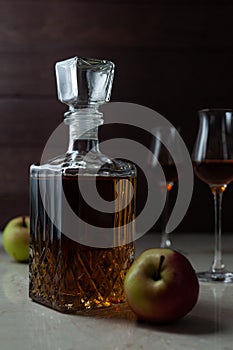 Vintage jar with homemade calvados with two apples and two glasses on marble table in dark mood.