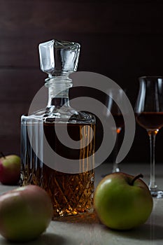 Vintage jar with homemade craft calvados with three apples and two glasses on marble table in dark mood.