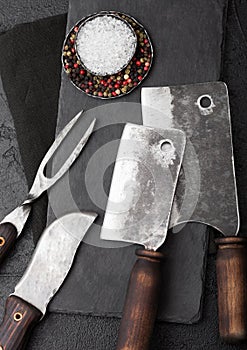 Vintage meat knife and fork and hatchets with stone chopping board and black table background. Butcher utensils. Close up